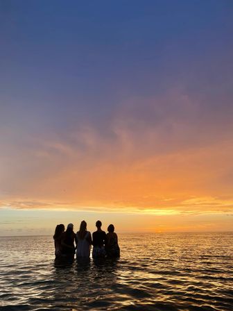 Five friends standing waist-deep in calm ocean water, silhouetted against a vivid orange and purple beach sunset over the horizon.