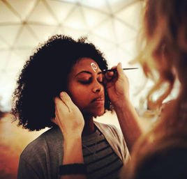 Person with eyes closed receiving a white swirl face paint above the eyebrow from an artist using a fine brush inside a geodesic dome — festival-style face-painting scene.