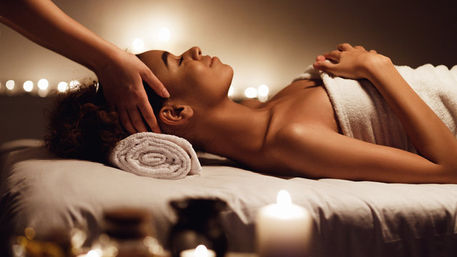 Candlelit spa head massage — relaxed woman wrapped in a towel lies on a massage table with a rolled towel under her head while a therapist’s hands give a soothing scalp treatment in warm ambient light