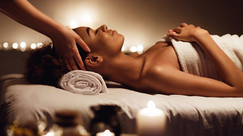 Candlelit spa head massage — relaxed woman wrapped in a towel lies on a massage table with a rolled towel under her head while a therapist’s hands give a soothing scalp treatment in warm ambient light