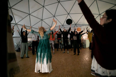 Leader in a green flowing dress guides a community with arms raised during an indoor movement workshop inside a geodesic dome.
