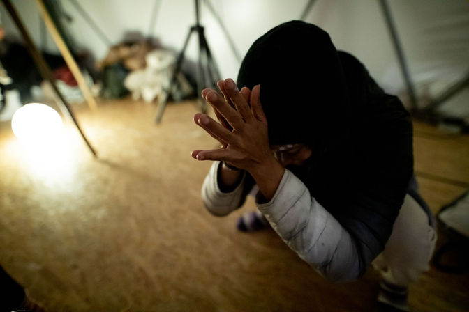 Low-light indoor scene of a hooded person crouching on a wooden floor, hands clasped over their face beside a glowing round lamp and tripod — moody studio-style portrait.