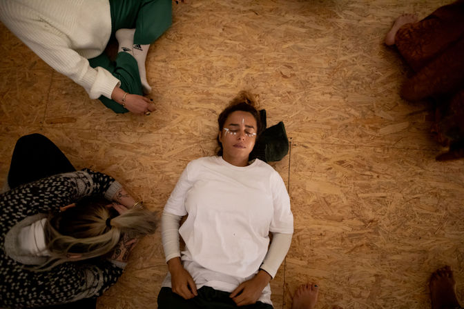 Participant lying on a plywood studio floor with cooling eye patches, eyes closed, surrounded by others sitting in a relaxed circle — calm group meditation and wellness workshop.