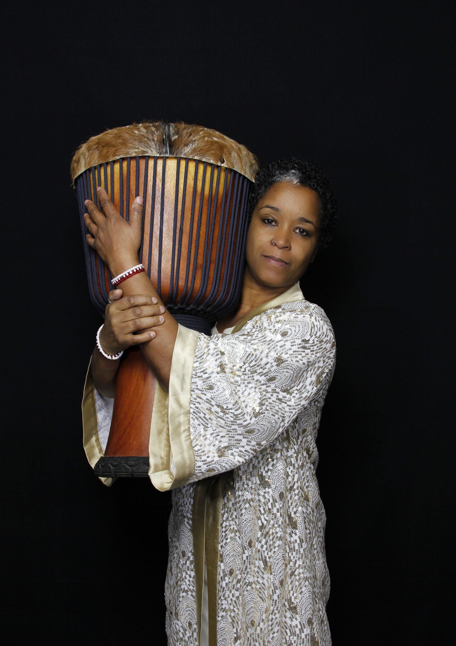 Studio portrait of a woman in a patterned cream and gold robe embracing a large wooden djembe drum with a fur head against a black background.