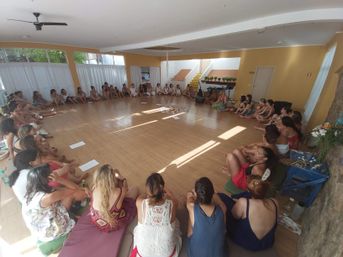 Sunlit indoor wellness studio with participants seated in a large circle on cushions around a central flower-and-candle arrangement, a bright wooden-floor group retreat or workshop setting.