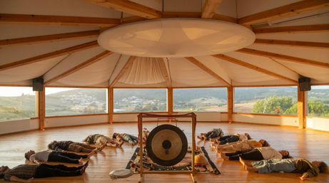 Sound bath meditation in a circular wooden studio with panoramic windows overlooking rolling hills; participants lie on the floor around a central gong at golden hour.