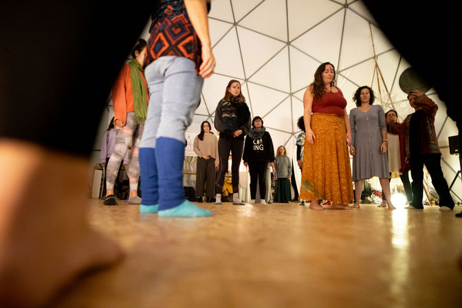 Barefoot adults standing in a community movement circle inside a white geodesic dome, wearing colorful casual clothes during a workshop.