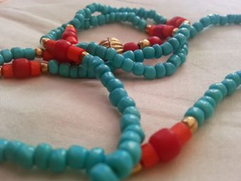 Close-up of turquoise seed-bead necklace with coral-red beads and small gold spacers laid on white fabric — boho-style summer jewelry.