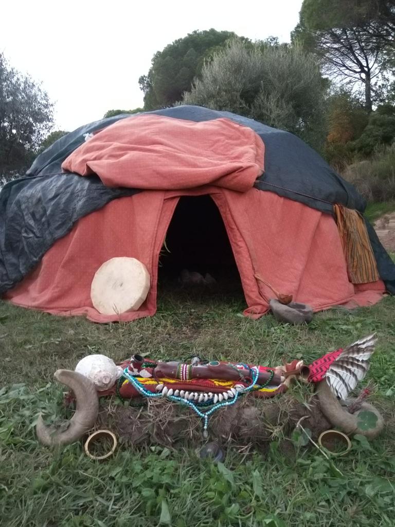 Orange dome tent in a grassy wooded clearing with a low ritual altar in front — hand drum, beads, shells, horns and feathers.