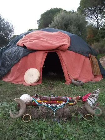 Orange dome tent in a grassy wooded clearing with a low ritual altar in front — hand drum, beads, shells, horns and feathers.