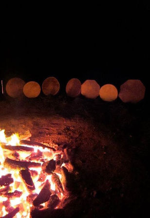 Outdoor nighttime campfire with glowing red-orange embers in the foreground and a row of blurred amber bokeh lights along the dark horizon