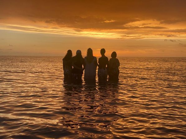 Five silhouetted figures standing waist‑deep in the ocean, watching a golden orange sunset with warm reflective ripples across calm water.