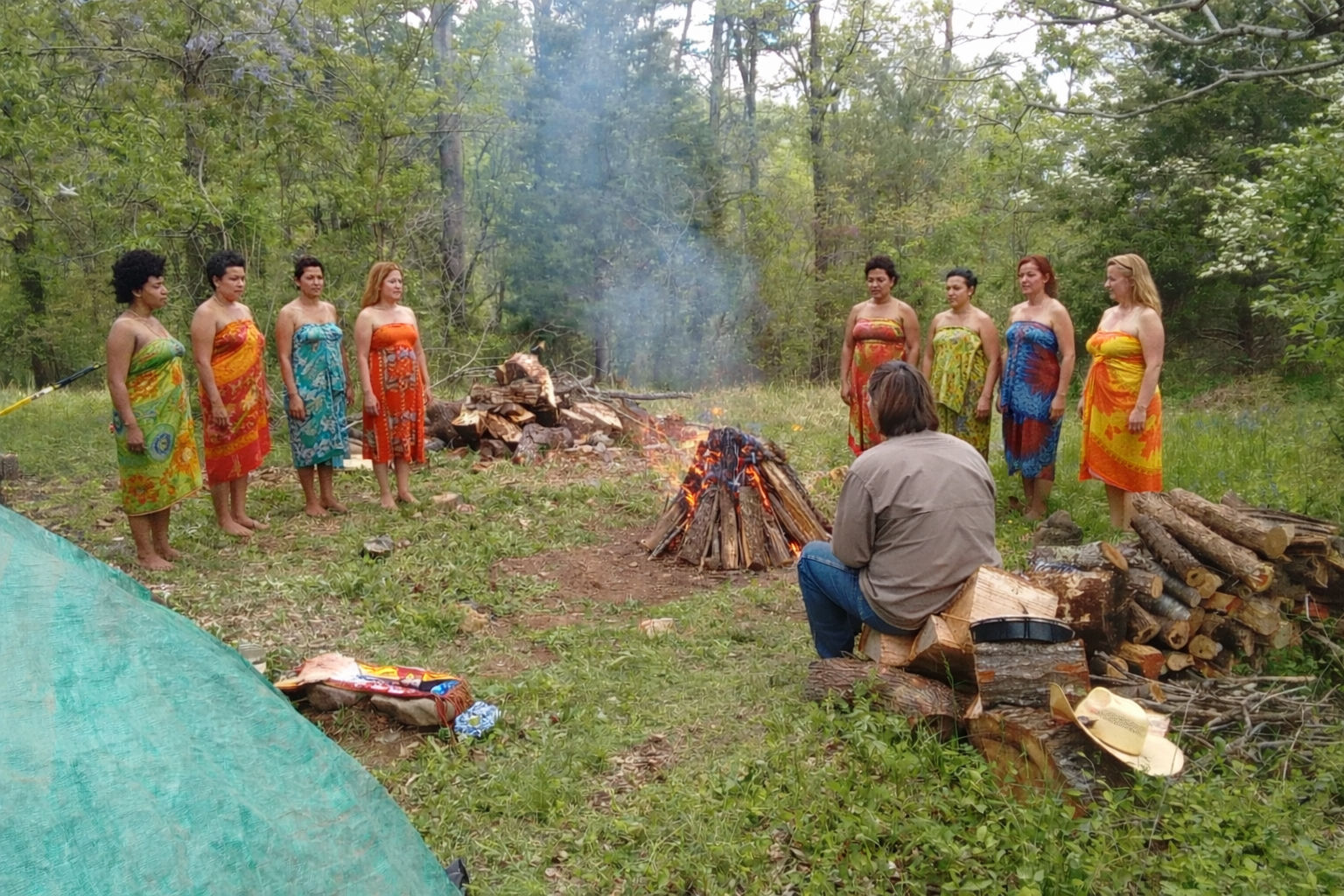 Group of women in colorful sarongs standing in a semicircle around a campfire in a wooded clearing while a person sits on stacked logs nearby, with firewood piles and camping gear on the grass.