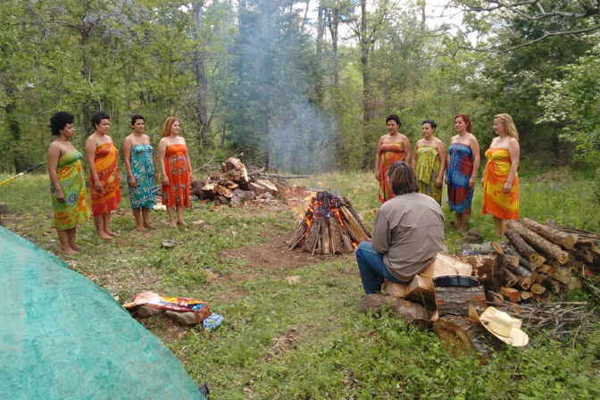 Group of women in colorful sarongs standing in a semicircle around a campfire in a wooded clearing while a person sits on stacked logs nearby, with firewood piles and camping gear on the grass.