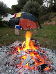 Cozy glowing campfire with bright embers in the foreground and a colorful fabric-covered dome tent on a grassy campsite backed by trees and a wooded hillside.