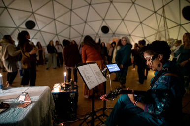 Candlelit community music circle inside a geodesic dome — seated guitarist with headset and face paint plays from sheet music while a group stands in a loose circle.