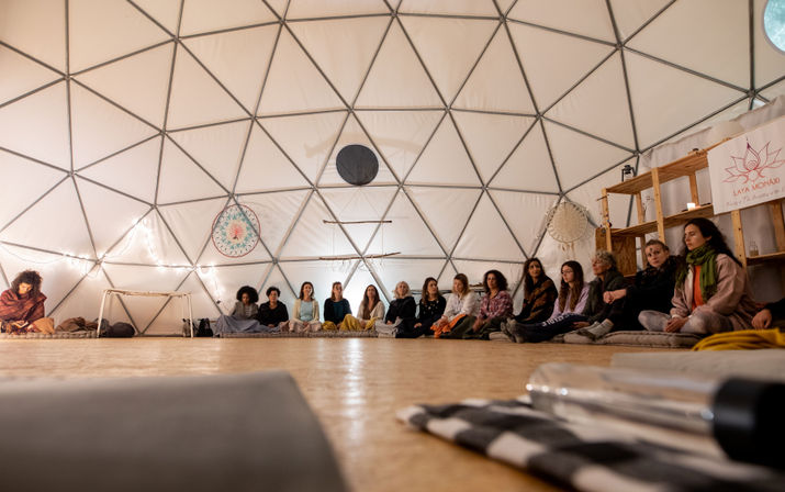 Cozy geodesic dome interior with a group of participants seated in a circle on cushions, string lights, dreamcatchers and wooden shelving creating a warm meditation/retreat atmosphere.