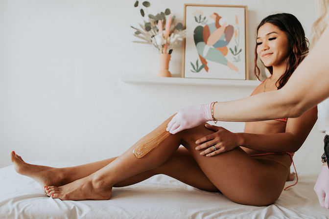 Smiling woman in a bikini receiving leg waxing as a technician in pink gloves applies warm wax on her lower leg in a bright, spa-style room with abstract wall art and a vase.