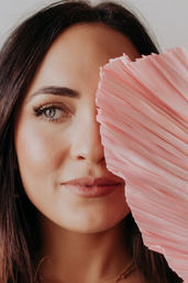 Close-up portrait of a smiling woman peeking from behind a pink pleated fan, showing one blue-gray eye, soft makeup and long dark hair.
