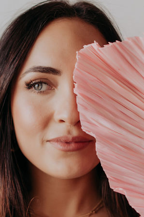 Close-up portrait of a smiling woman peeking from behind a pink pleated fan, showing one blue-gray eye, soft makeup and long dark hair.