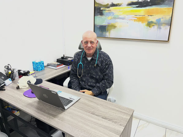 Friendly clinician with stethoscope seated at a modern clinic desk with a laptop, tissue box, paperwork and abstract wall art — bright medical office consultation scene.