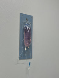 Pink-tinted 0.9% sodium chloride IV bag with tubing and drip chamber hanging on a blue wall mount in a bright medical clinic room