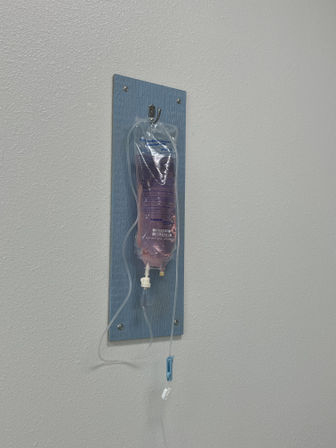 Pink-tinted 0.9% sodium chloride IV bag with tubing and drip chamber hanging on a blue wall mount in a bright medical clinic room