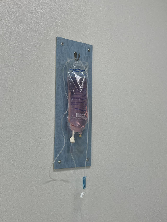Pink-tinted 0.9% sodium chloride IV bag with tubing and drip chamber hanging on a blue wall mount in a bright medical clinic room