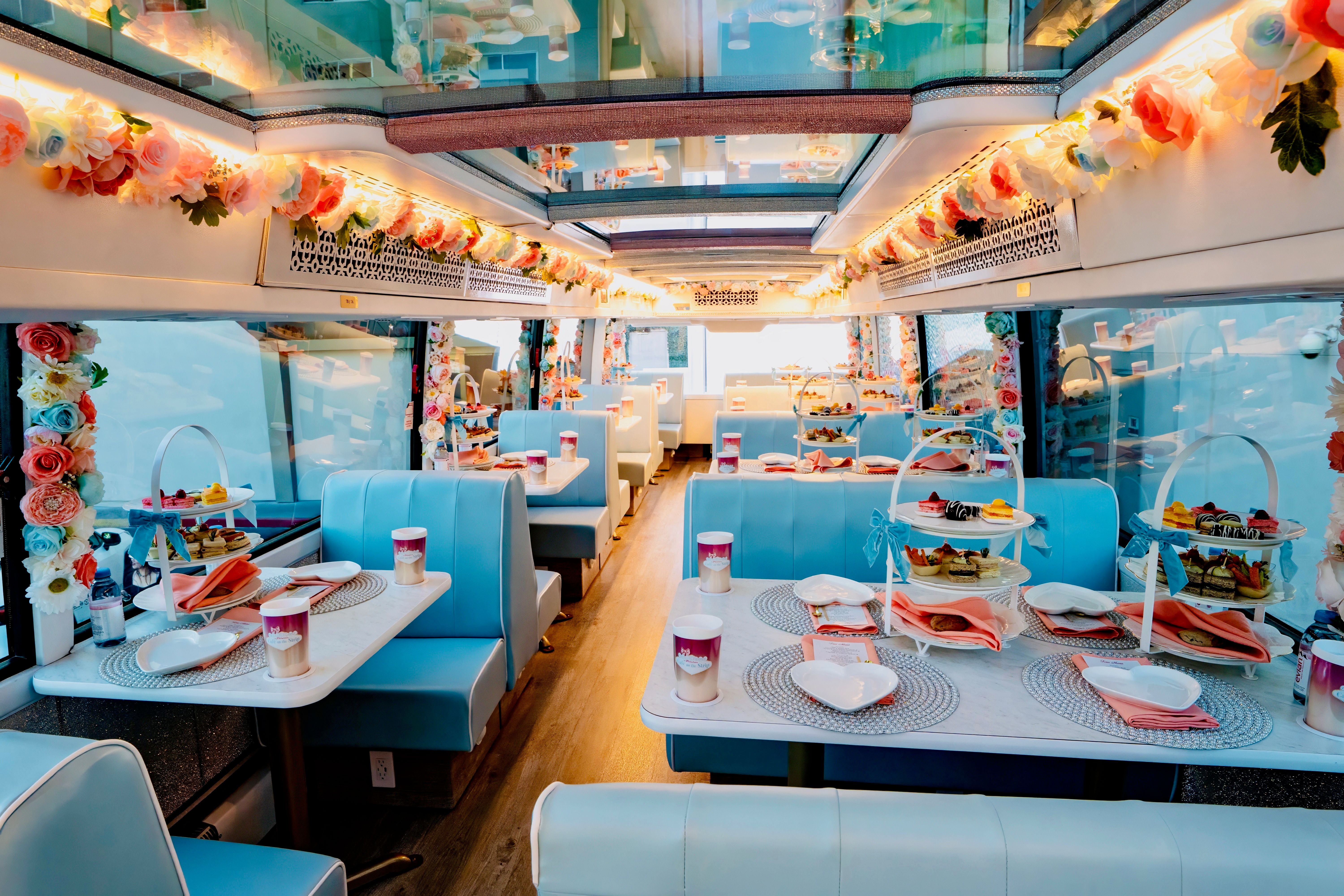 Pastel-blue retro-style party bus interior set for high tea with floral garlands, booth seating and tiered pastry stands