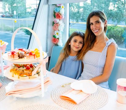 Woman and young girl smiling during a pastel-themed afternoon tea in a windowed booth, with tiered trays of pastries and sandwiches, floral garland, pink napkins and heart-shaped plates.