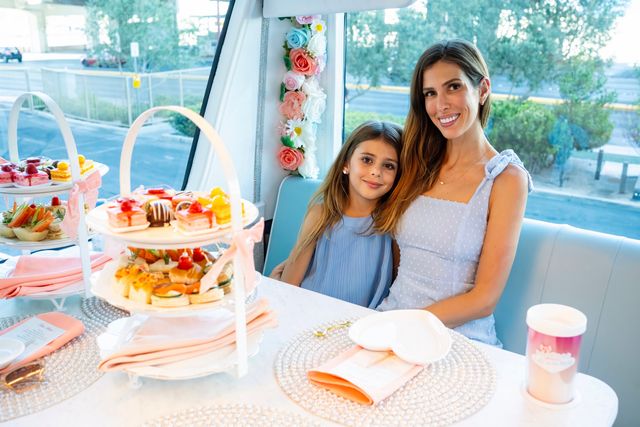 Woman and young girl smiling during a pastel-themed afternoon tea in a windowed booth, with tiered trays of pastries and sandwiches, floral garland, pink napkins and heart-shaped plates.
