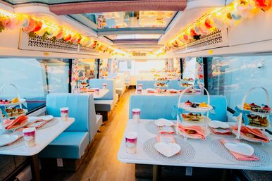 Bright pastel tea party inside a decorated train dining car with turquoise booth seating, skylights, floral garlands and string lights, marble-top tables set with tiered dessert stands, pink napkins and heart-shaped plates.