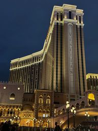 Night view of an illuminated Renaissance-inspired hotel tower and ornate Italianate arcade with arched bridge, glowing street lamps and silhouetted pedestrians on the Las Vegas Strip.