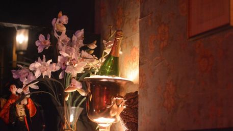 Champagne bottle chilling in a vintage silver ice bucket beside pink orchids and a porcelain figurine in a cozy, dimly lit corner with floral wallpaper.