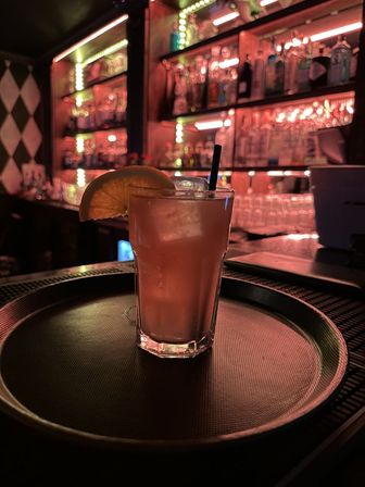 Short cocktail glass with ice, lemon wedge and straw on a round serving tray at a neon-lit bar with blurred backlit liquor shelves