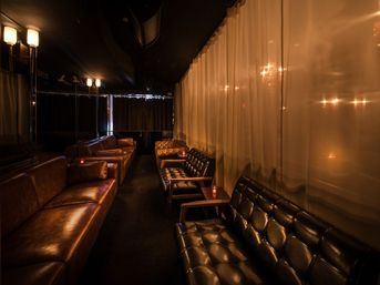 Cozy dimly lit lounge interior with tufted leather sofas and armchairs along a narrow room, candlelit side tables and sheer curtains creating a warm amber ambiance.