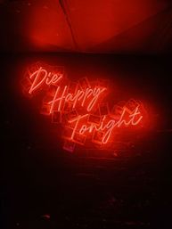 Red neon script sign reading 'Die Happy Tonight' mounted on a dark brick wall with scattered Polaroid photos and moody bar-style lighting.