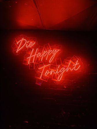 Red neon script sign reading 'Die Happy Tonight' mounted on a dark brick wall with scattered Polaroid photos and moody bar-style lighting.