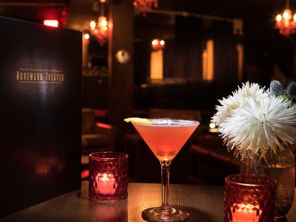 Dimly lit upscale cocktail lounge table with a pink martini garnished with a lemon twist, glowing red votive candles and a white chrysanthemum bouquet against a cozy, intimate bar backdrop.