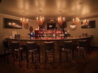Cozy dimly lit cocktail bar interior with a curved wooden counter, leather bar stools, backlit liquor shelves, wall-mounted TVs and ornate amber chandeliers casting warm reflections.