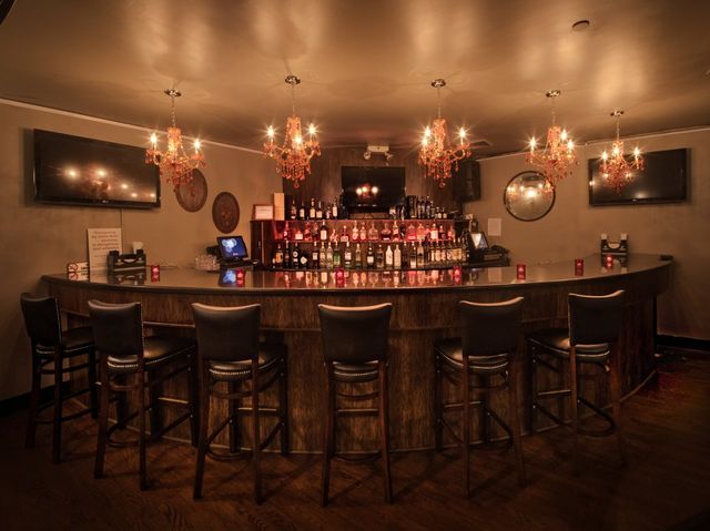 Cozy dimly lit cocktail bar interior with a curved wooden counter, leather bar stools, backlit liquor shelves, wall-mounted TVs and ornate amber chandeliers casting warm reflections.