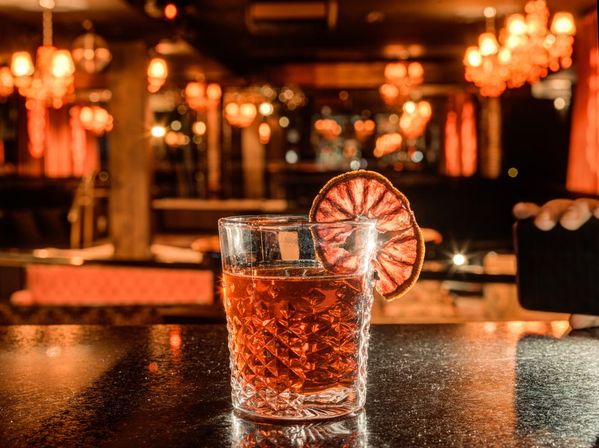 Amber cocktail in a faceted glass garnished with a dried orange slice, resting on a glossy bar counter in a warm, dimly lit lounge with chandeliers and bokeh lights.
