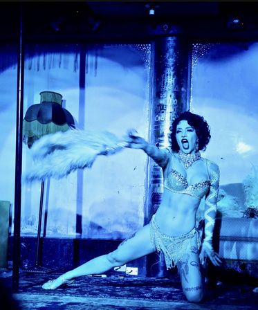 Burlesque performer on a cabaret stage bathed in blue light, wearing a sparkling fringe costume and jeweled bra, kneeling in a dramatic pose while waving a large feather fan.