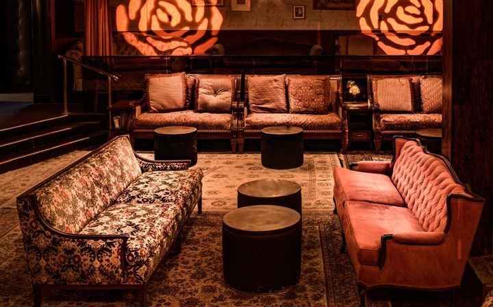 Cozy vintage lounge interior with tufted velvet sofa and floral-pattern settee facing round low tables, warm amber lighting and rose-shaped wall light projections over patterned rugs