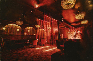 Red-lit retro lounge interior with crystal chandeliers, velvet armchairs, small tables and vertical string curtains creating a sultry cabaret vibe.