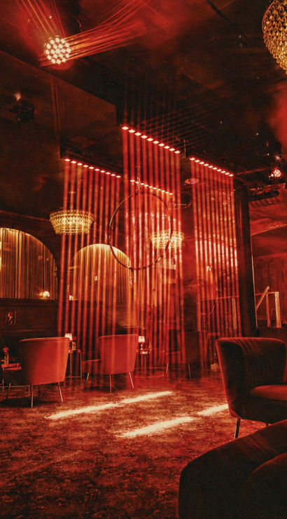 Red-lit retro lounge interior with crystal chandeliers, velvet armchairs, small tables and vertical string curtains creating a sultry cabaret vibe.