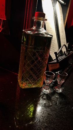 Crystal-cut decanter of amber liquor and two small shot glasses on a dimly lit bar countertop with red ambient lighting and a menu in the background.