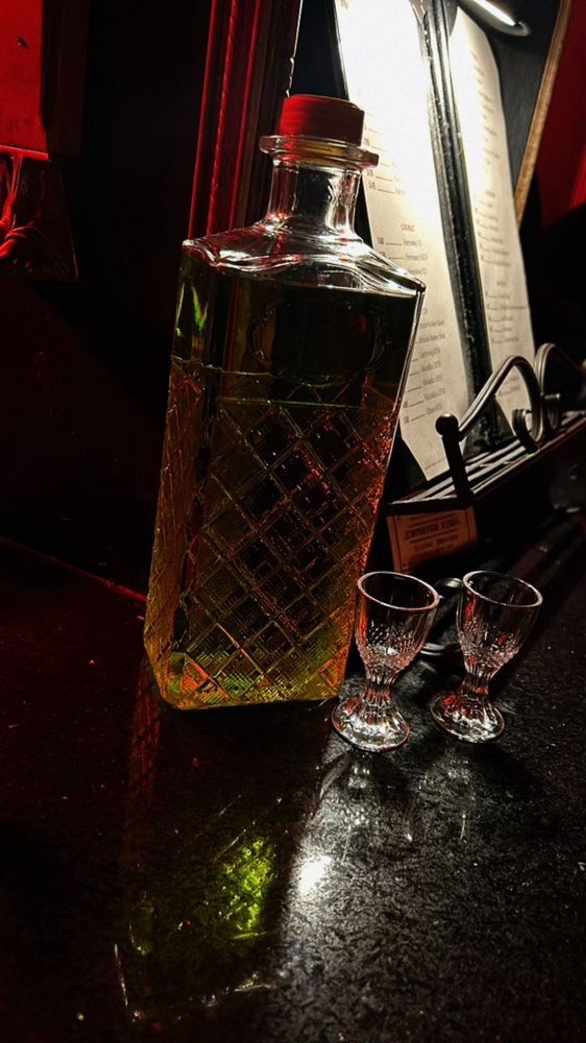 Crystal-cut decanter of amber liquor and two small shot glasses on a dimly lit bar countertop with red ambient lighting and a menu in the background.