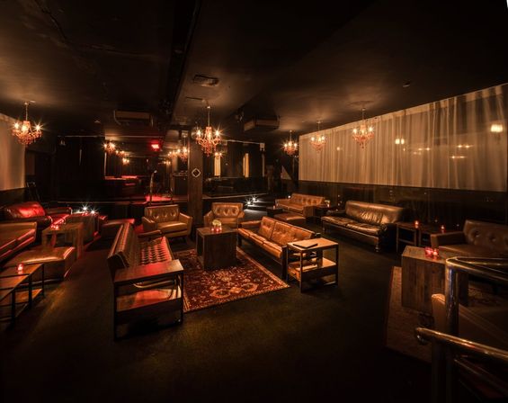 Dimly lit urban lounge interior with leather sofas and armchairs around wooden coffee tables and rugs, warm amber chandeliers and candles, intimate cocktail bar vibe with a small raised stage in the background.
