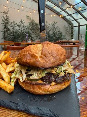 Close-up gourmet burger on a toasted brioche bun topped with slaw, pickles and sauce, skewered by a knife and served with seasoned fries on a slate board in a string-lit outdoor patio.